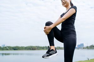  Person in black athletic wear performing knee mobility exercise near lake with city skyline background