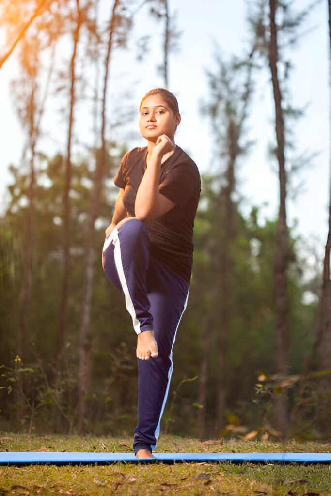 Person performing single-leg balance exercise wearing black t-shirt and navy track pants on exercise mat in forest setting during golden hour