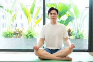 Person meditating in white t-shirt on yoga mat by window with tropical plants, demonstrating peaceful recovery environment