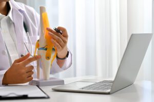 Healthcare provider in clinical setting analyzing knee joint model at desk with laptop computer