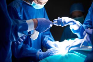 Surgeons in blue scrubs and protective gear performing surgery with bright surgical lights illuminating the operating field.