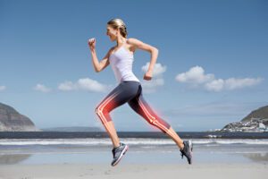 Athletic person running on beach wearing white tank top and gray leggings, with skeletal overlay showing knee joint mechanics